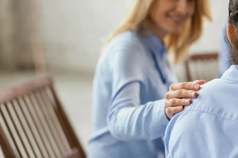 A supportive gesture in a therapy or support group setting, showing one woman gently placing her hand on another’s shoulder, symbolizing empathy and connection.