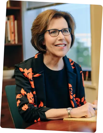 Smiling woman in floral blazer sits at a desk with a notepad.