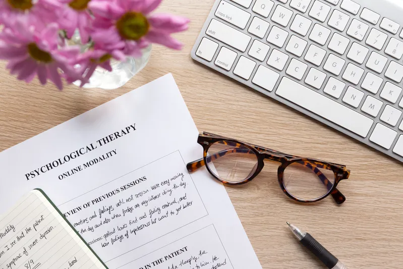 Online psychological therapy paperwork on desk with keyboard, glasses, and clinical session notes
